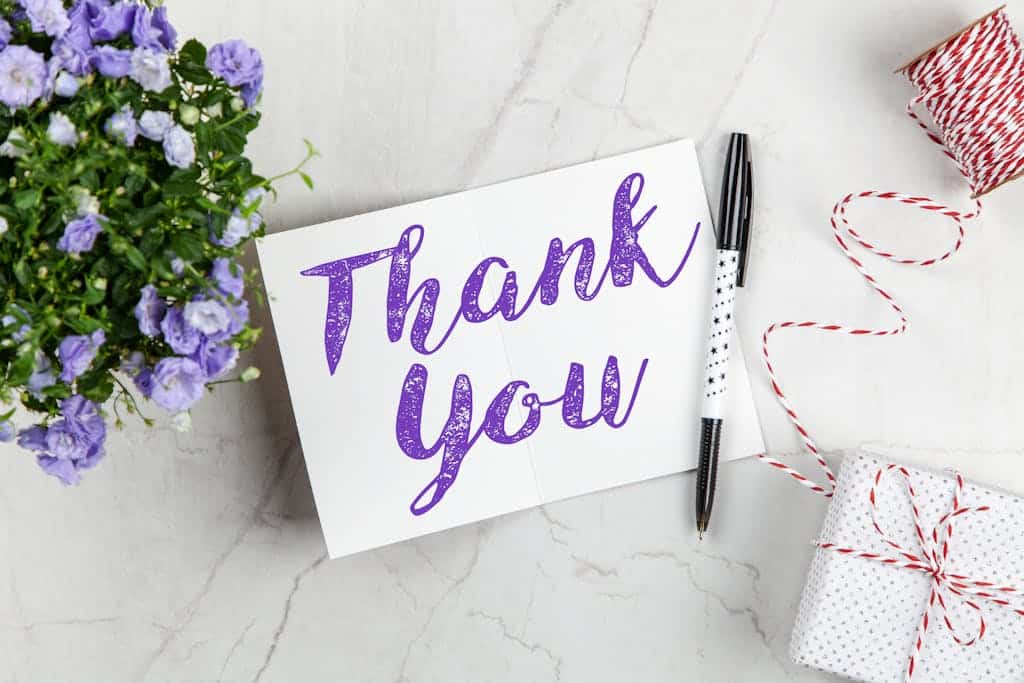 A stylish thank you card paired with purple flowers, a gift box, and pen on a marble surface.